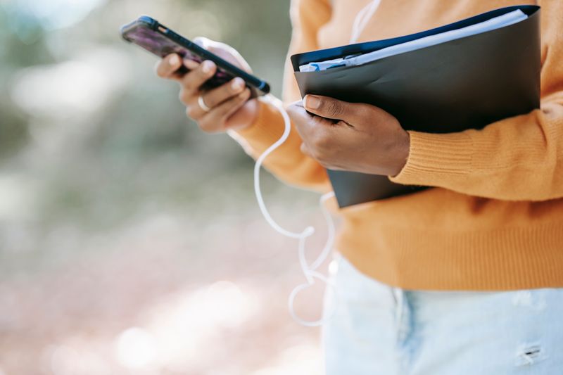 A Black woman checking her smartphone outdoors with work documents in hand